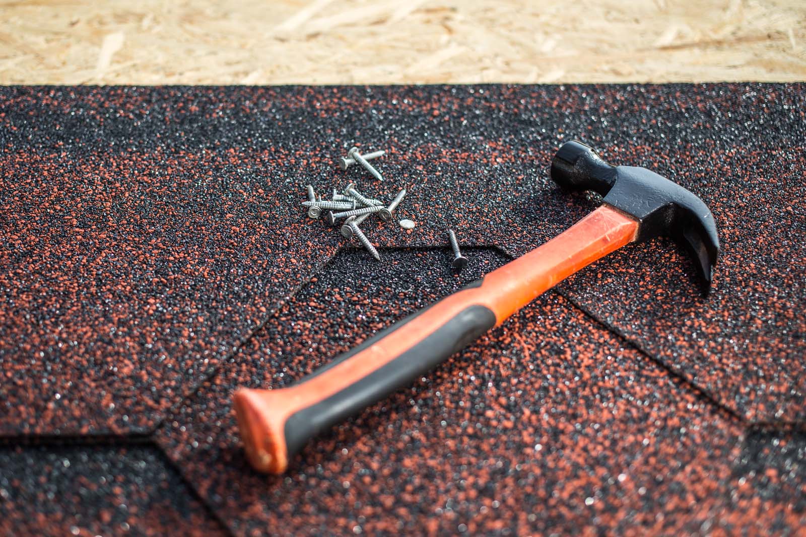 A hammer and several nails are placed on top of red and black asphalt shingles, which a roofing contractor might use, all set on a wooden surface.